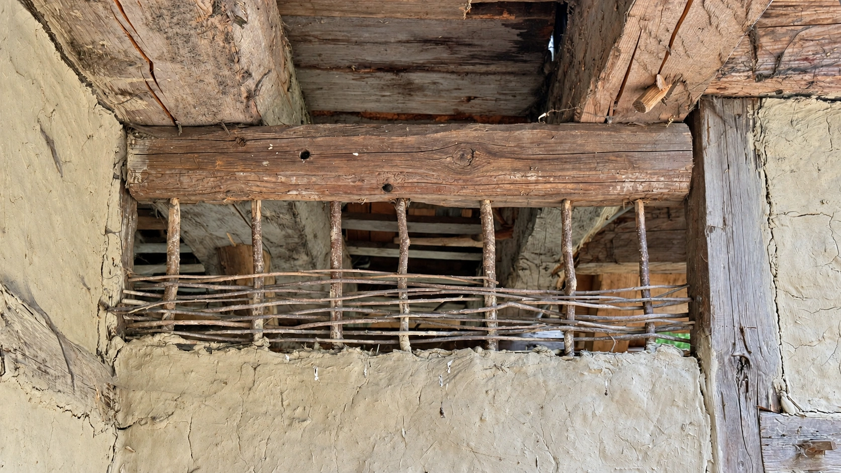 Vertical stakes support woven rods within the timber frame. The exposed section reveals the wattle beneath the daub.